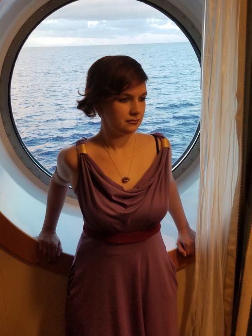 Caitlin in a purple dress standing in front of a window that looks out to open waters