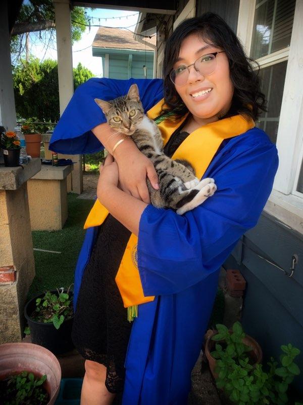 clara in yellow and blue grad attire and a black dress holding a cat smiling