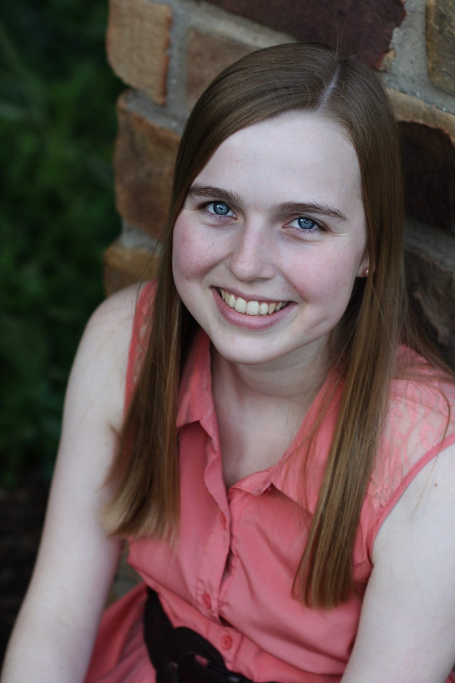 elizabeth smiling in a pink shirt sitting against a brick wall and shrubbery