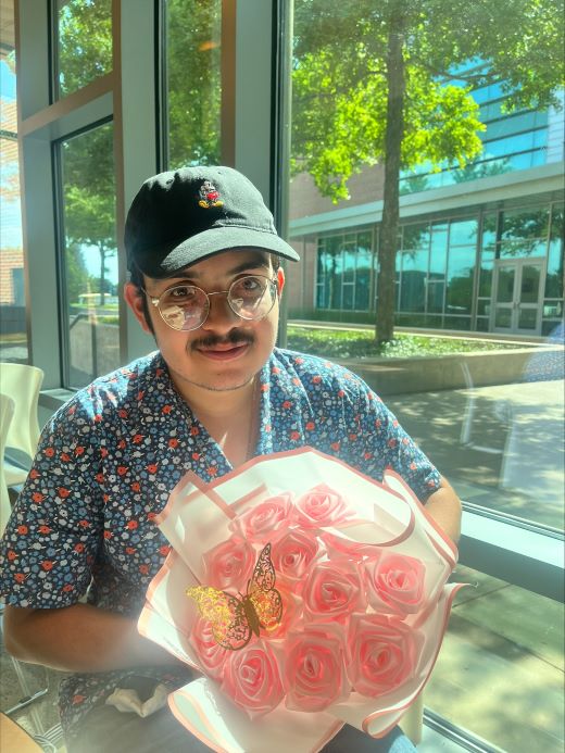 jose in a ball cap and glasses beside a window with sunlight streaming in, handmade pink flowers in the bottom right corner